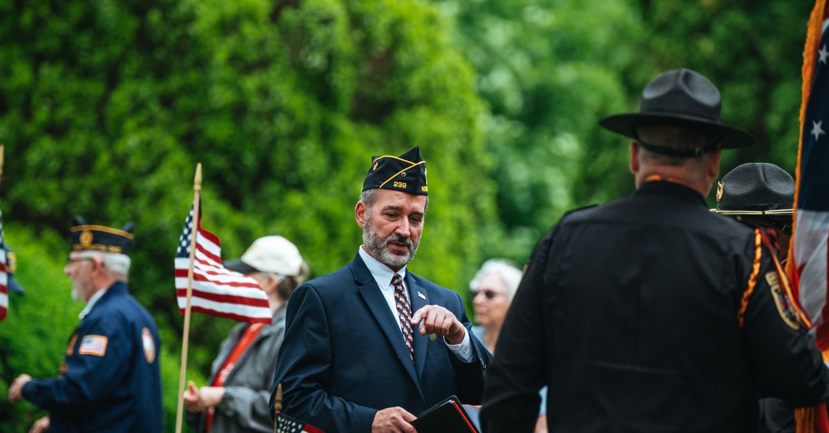 Veterans gathered in formal attire holding American flags during a memorial ceremony outdoors.